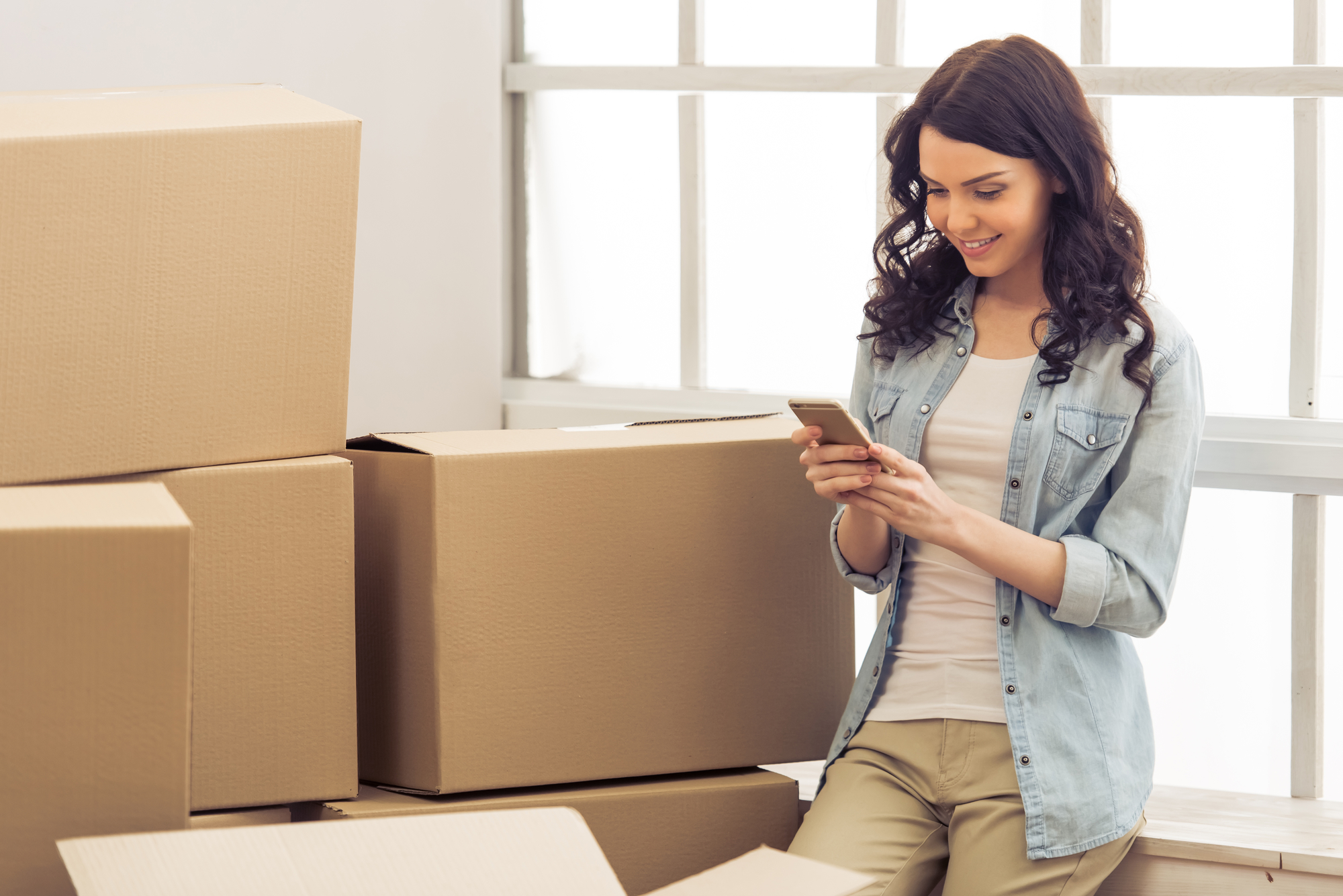 Young woman moving, standing among cardboard boxes and using a smartphone