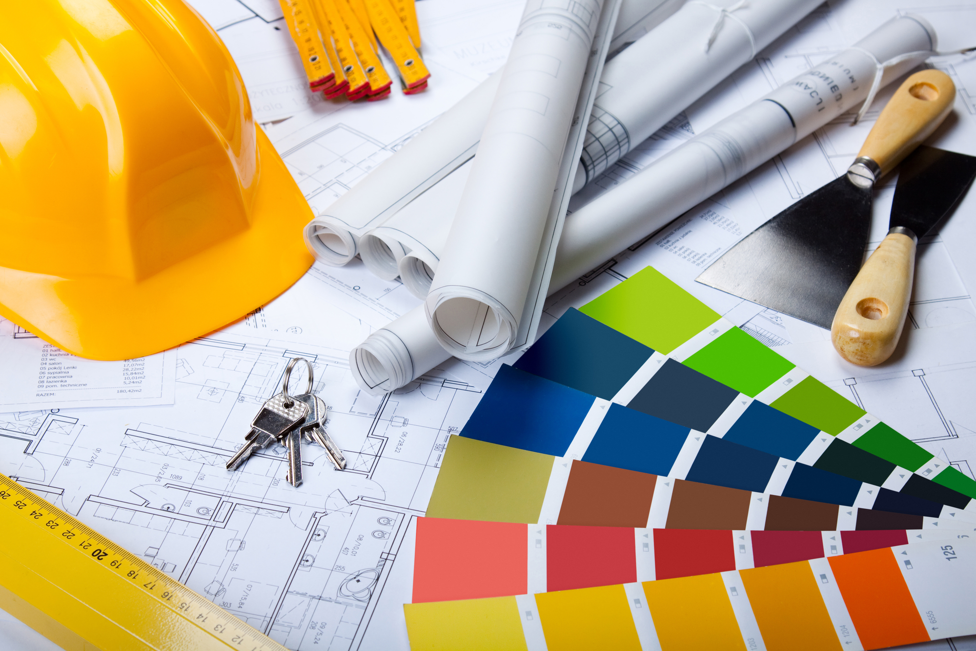 Construction plans with rolled blueprints, a yellow hard hat, paint color swatches, keys, and construction tools on a table.